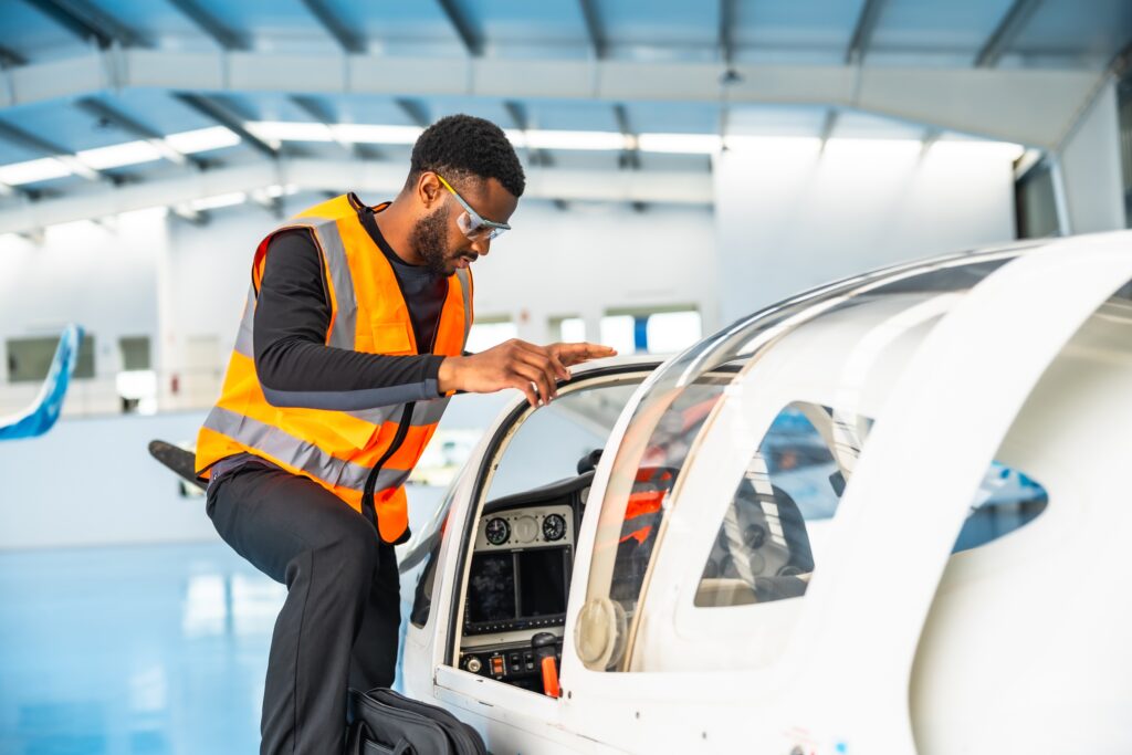 Maintenance technician in a high-visibility vest and safety glasses inspecting the door frame mechanism of a light aircraft in a hangar.