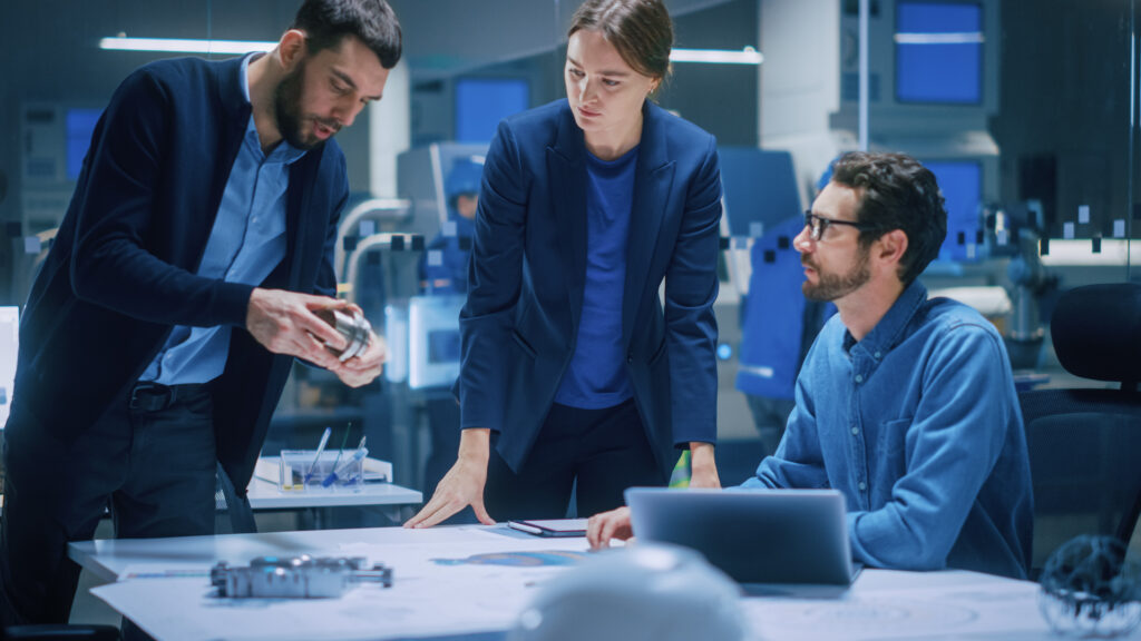 Üç mühendis modern bir Ar-Ge tesisinde iş birliği yapıyor, bir adam işlenmiş bir metal parçayı elinde tutuyor.