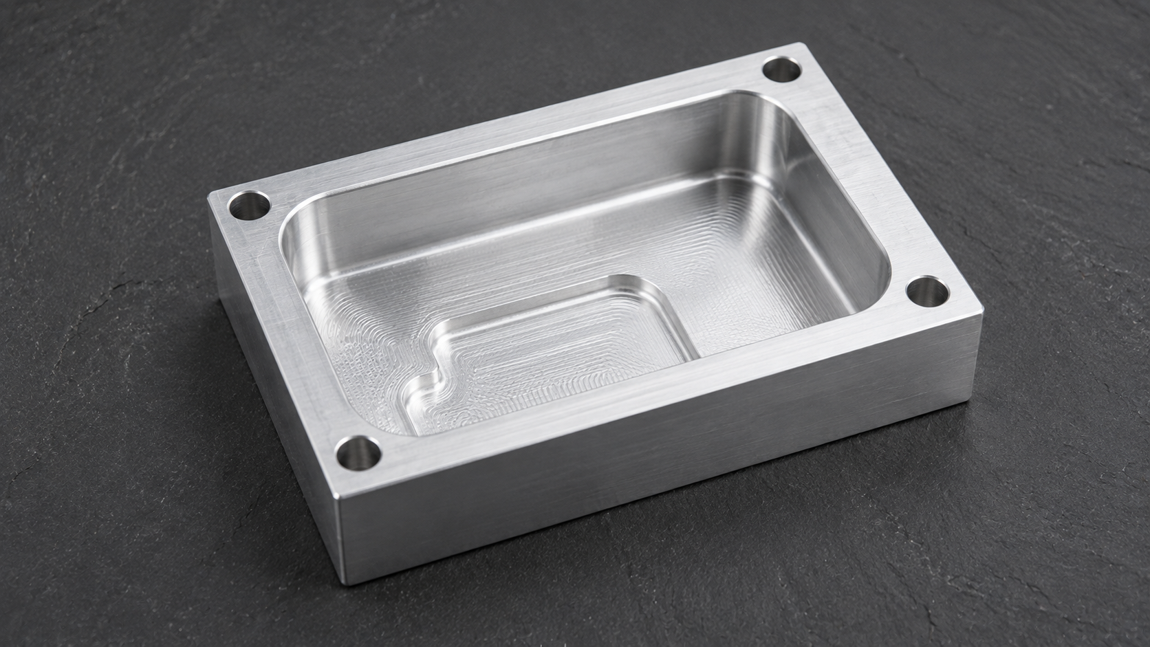 Machined aluminum block with a deep rectangular pocket and filleted internal corners on a matte slate surface.